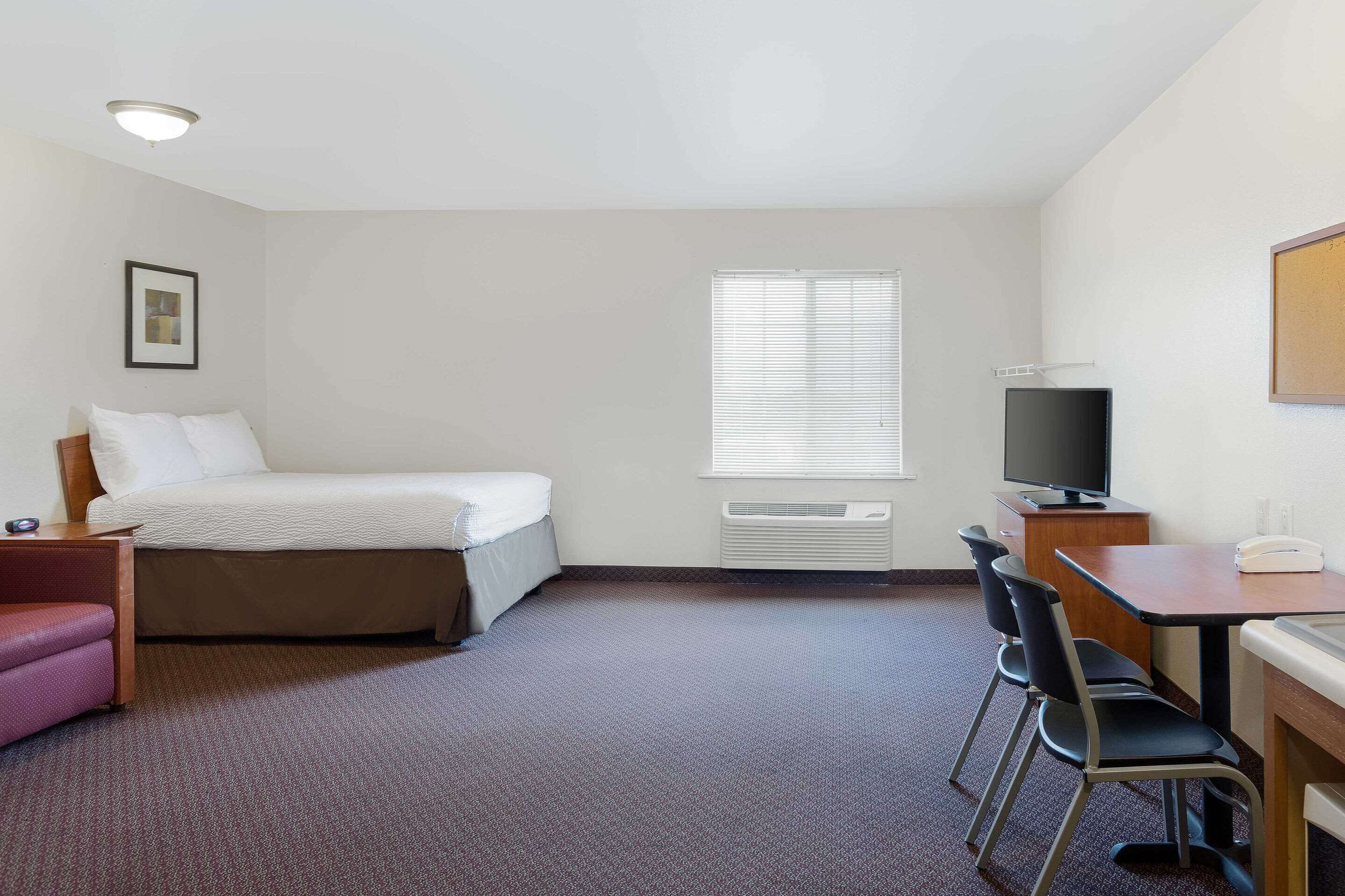 Habitación de hotel con cama, escritorio, silla, televisor y ventana con persianas.
