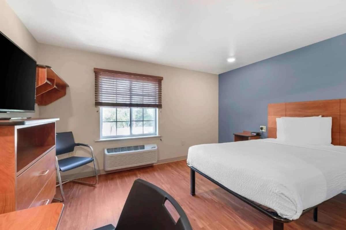 A hotel room with a bed, a desk with a chair, a television, a window with blinds, and a heater.
