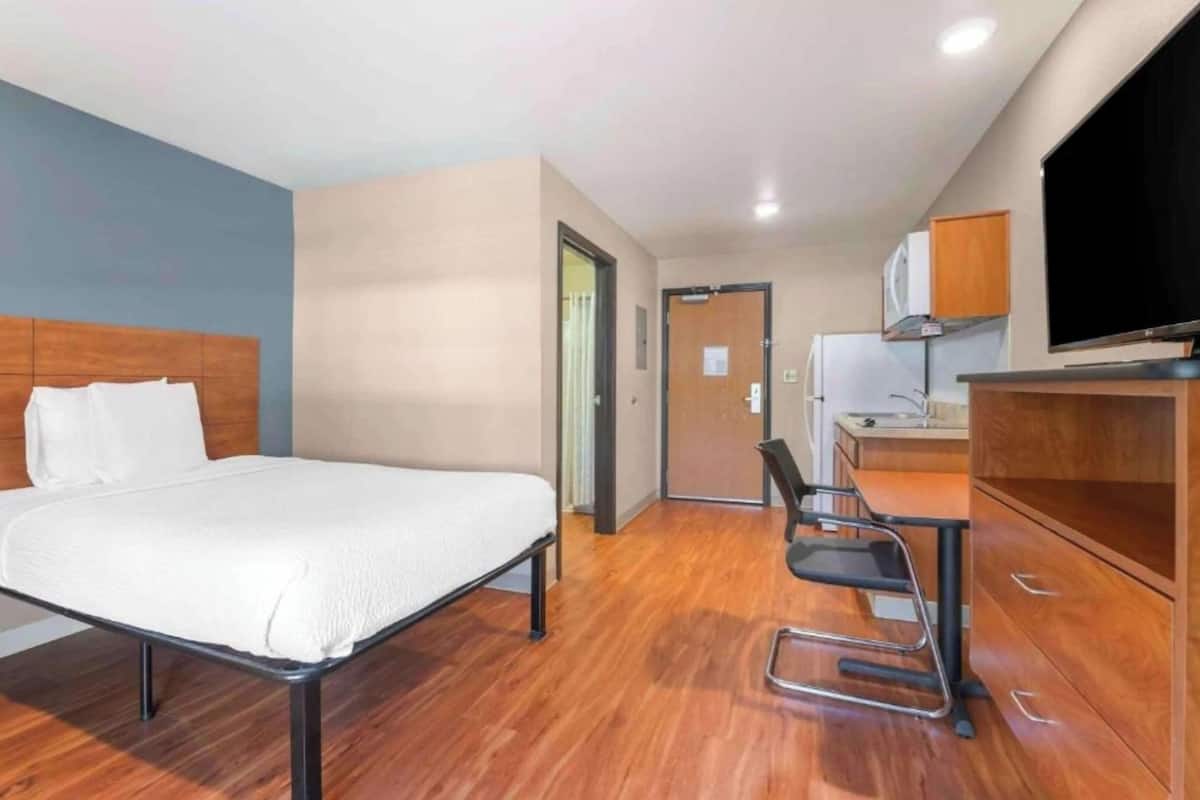 A hotel room with a bed, a desk, a chair, a television, and a kitchenette.