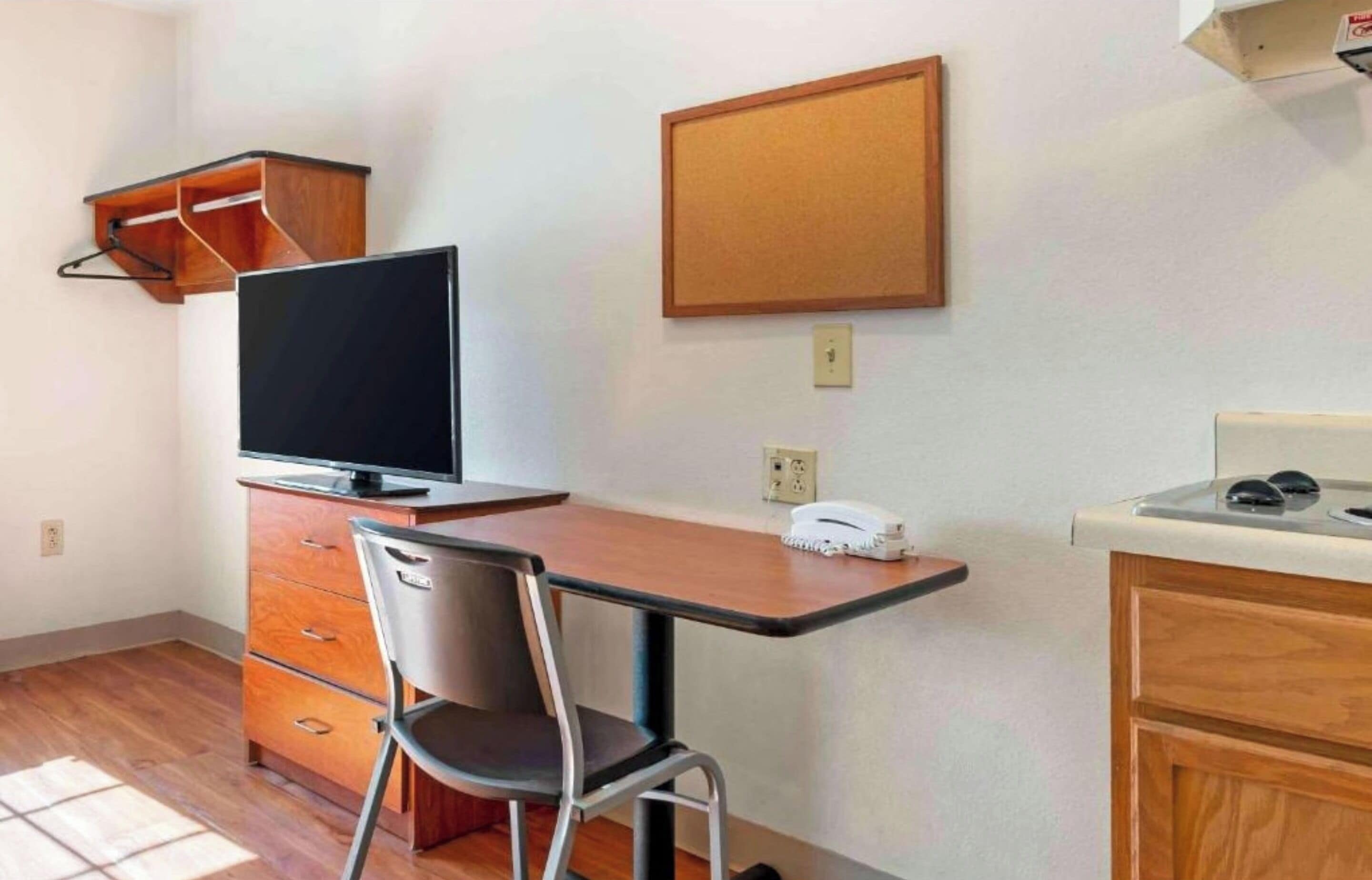 A small room with a wooden desk, a chair, a television, a corkboard, and a telephone.