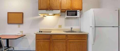 A compact kitchen with wooden cabinets, a microwave, a refrigerator, a small table, and a chair.
