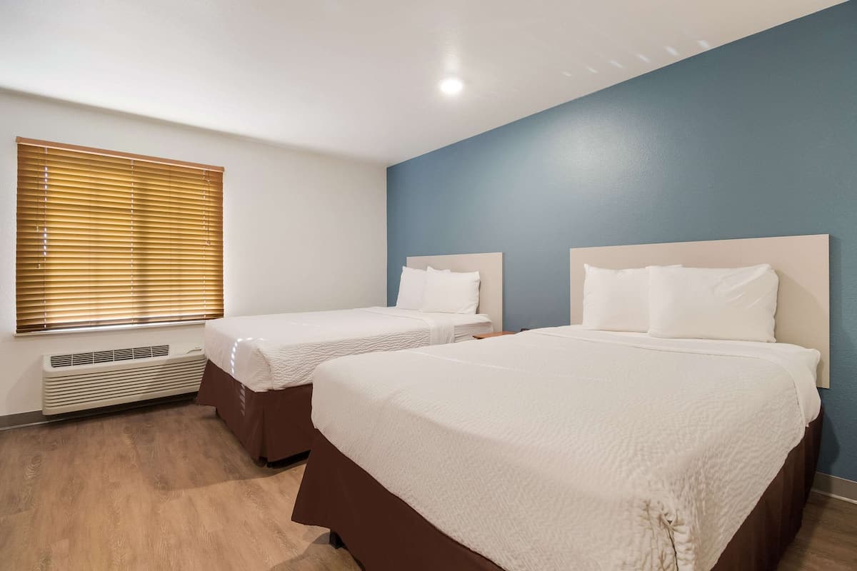A hotel room with two beds, a window with blinds, a wall heater, and a blue accent wall.
