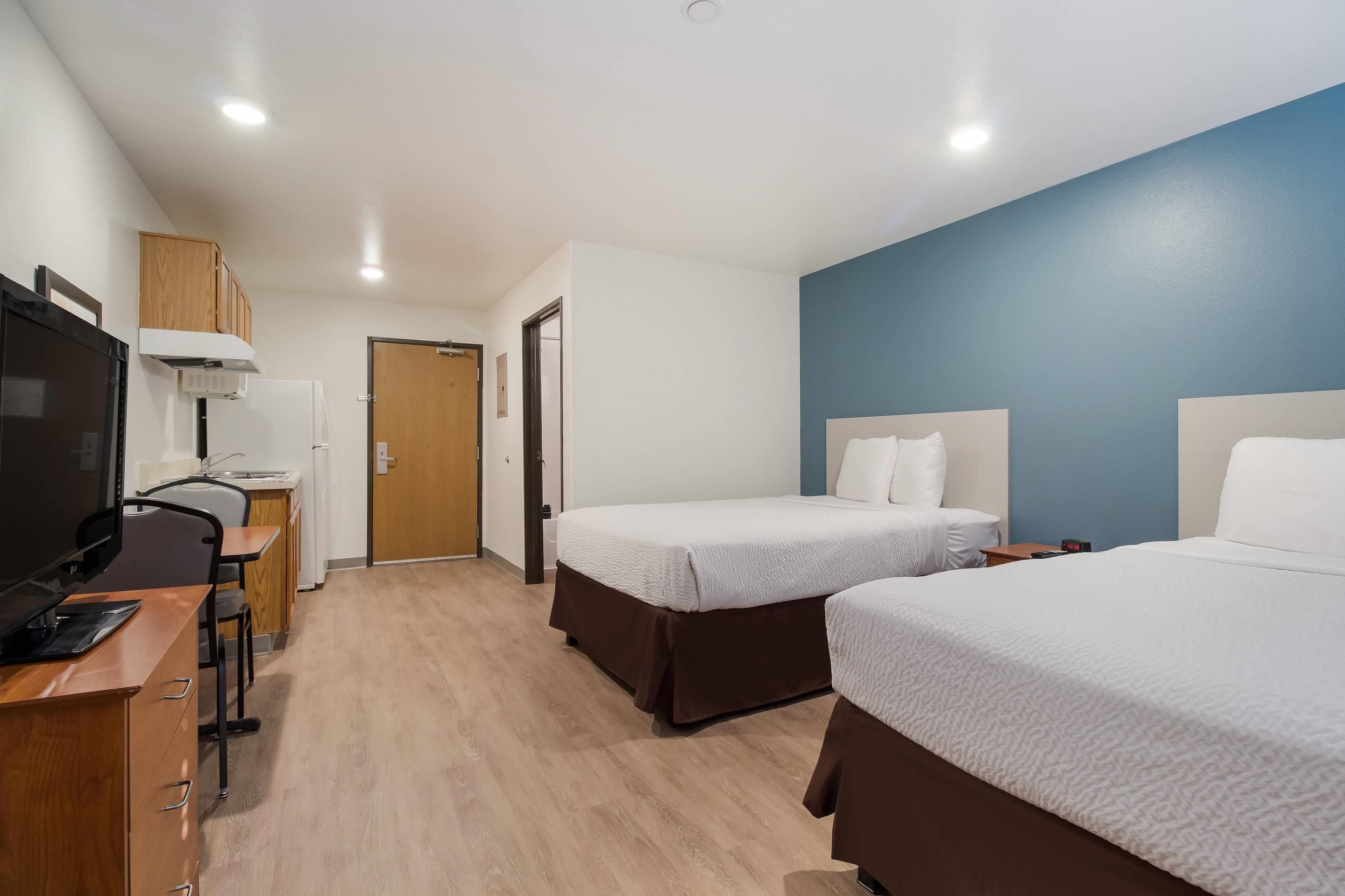Una habitación de hotel con dos camas, un escritorio con computadora, televisor y kitchenette.