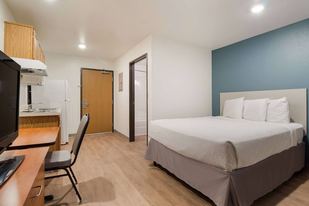 A hotel room with a bed, desk, chair, and kitchenette.