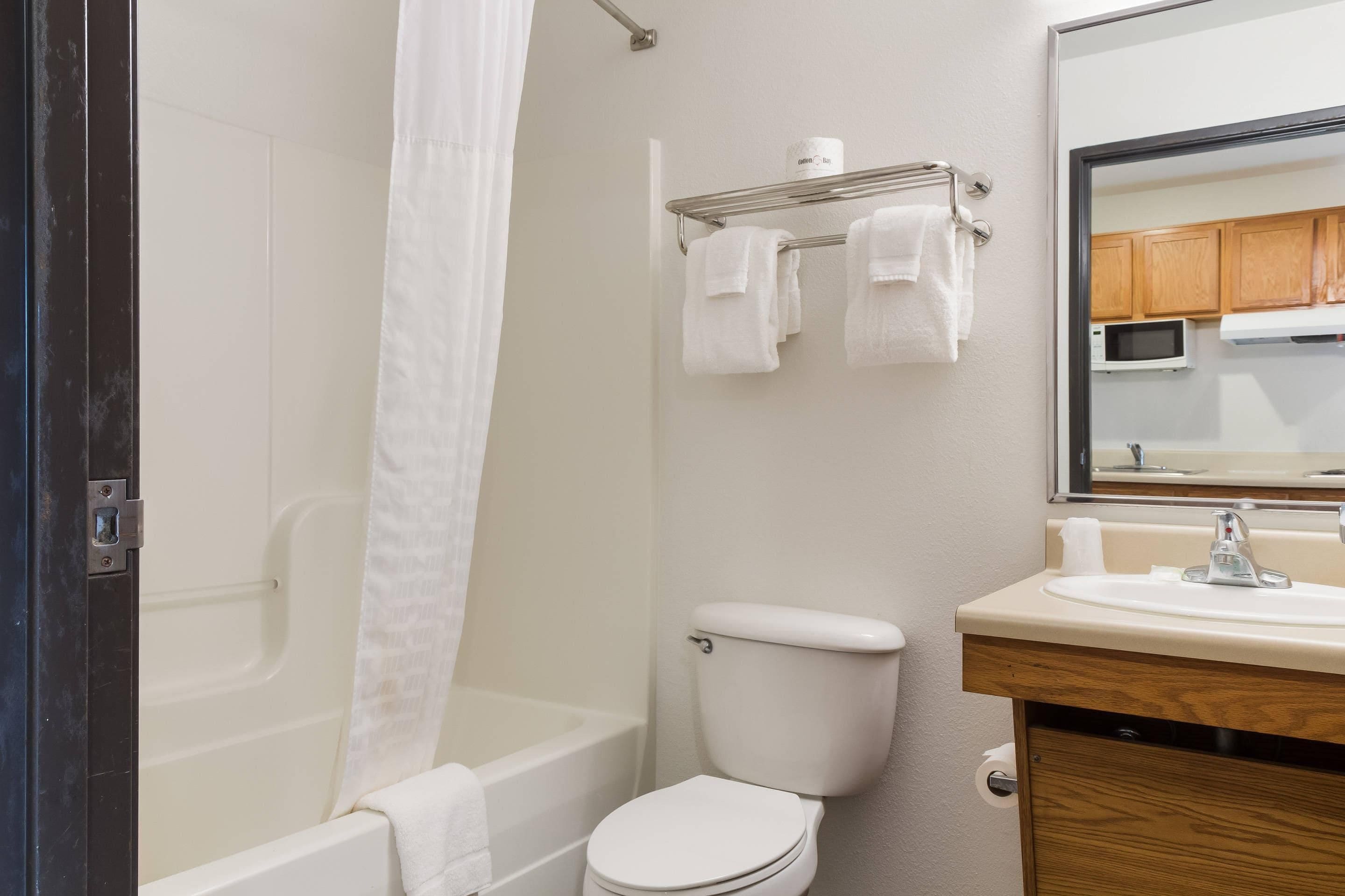 A bathroom with a bathtub, toilet, sink, and a mirror reflecting a kitchen area.