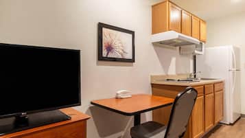 A compact kitchenette with a flat-screen TV, a small table, a chair, wooden cabinets, a microwave, and a refrigerator.