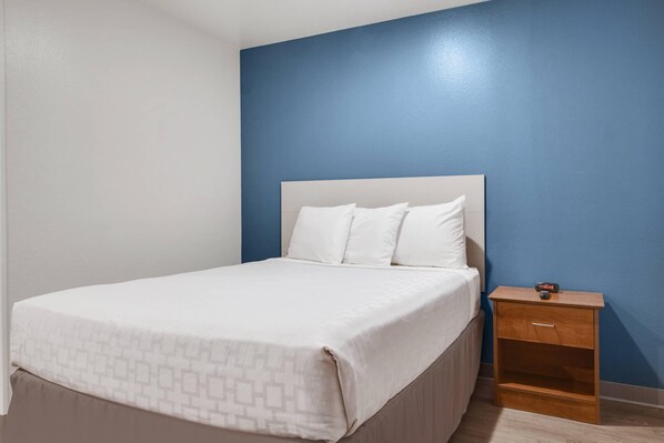 A single bed with a white headboard and pillows, a wooden nightstand with a drawer, and a blue wall.