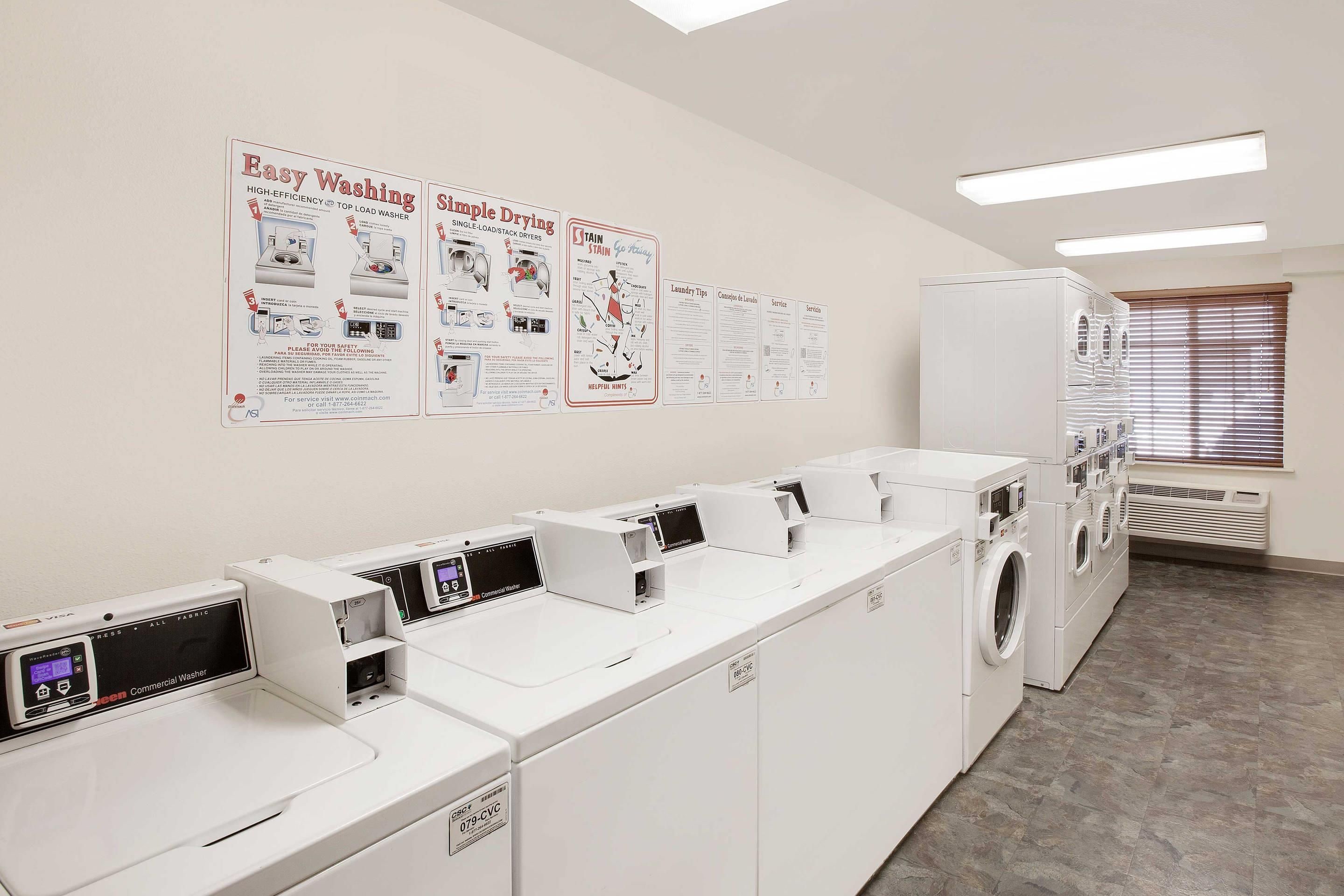 A laundry facility with multiple washing machines and dryers, instructional posters on the wall, and a window with blinds.