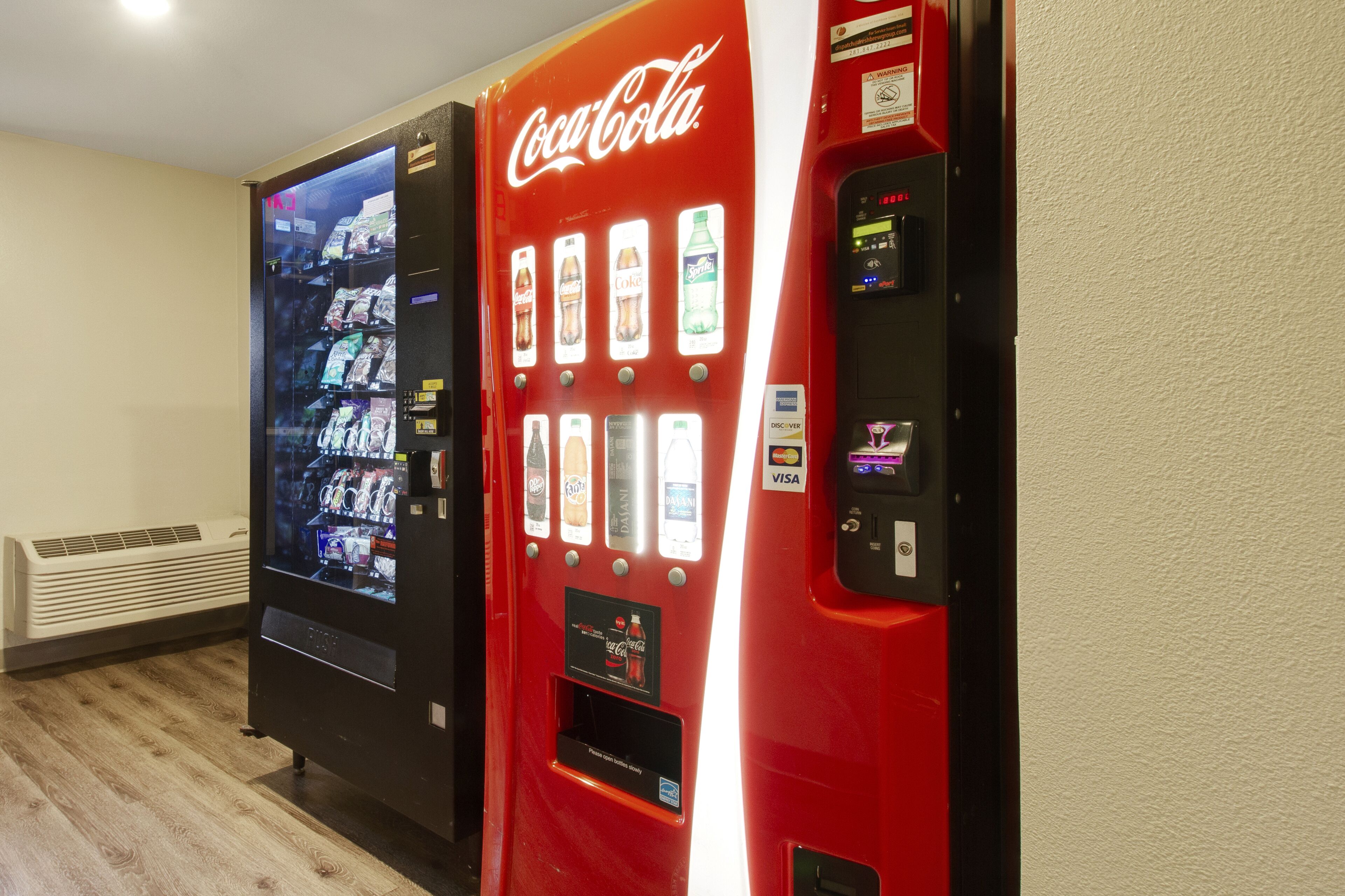 Two vending machines, one Coca-Cola and one Doritos, in a room with a white wall and a vent.