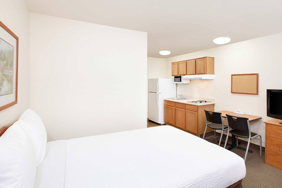 A hotel room with a bed, a desk with a chair, a microwave, a refrigerator, and wooden cabinets.