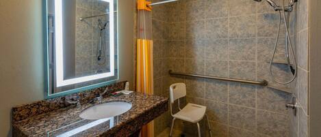 Un baño con lavabo de granito, un espejo, ducha con barra de apoyo y asiento accesible para silla de ruedas.