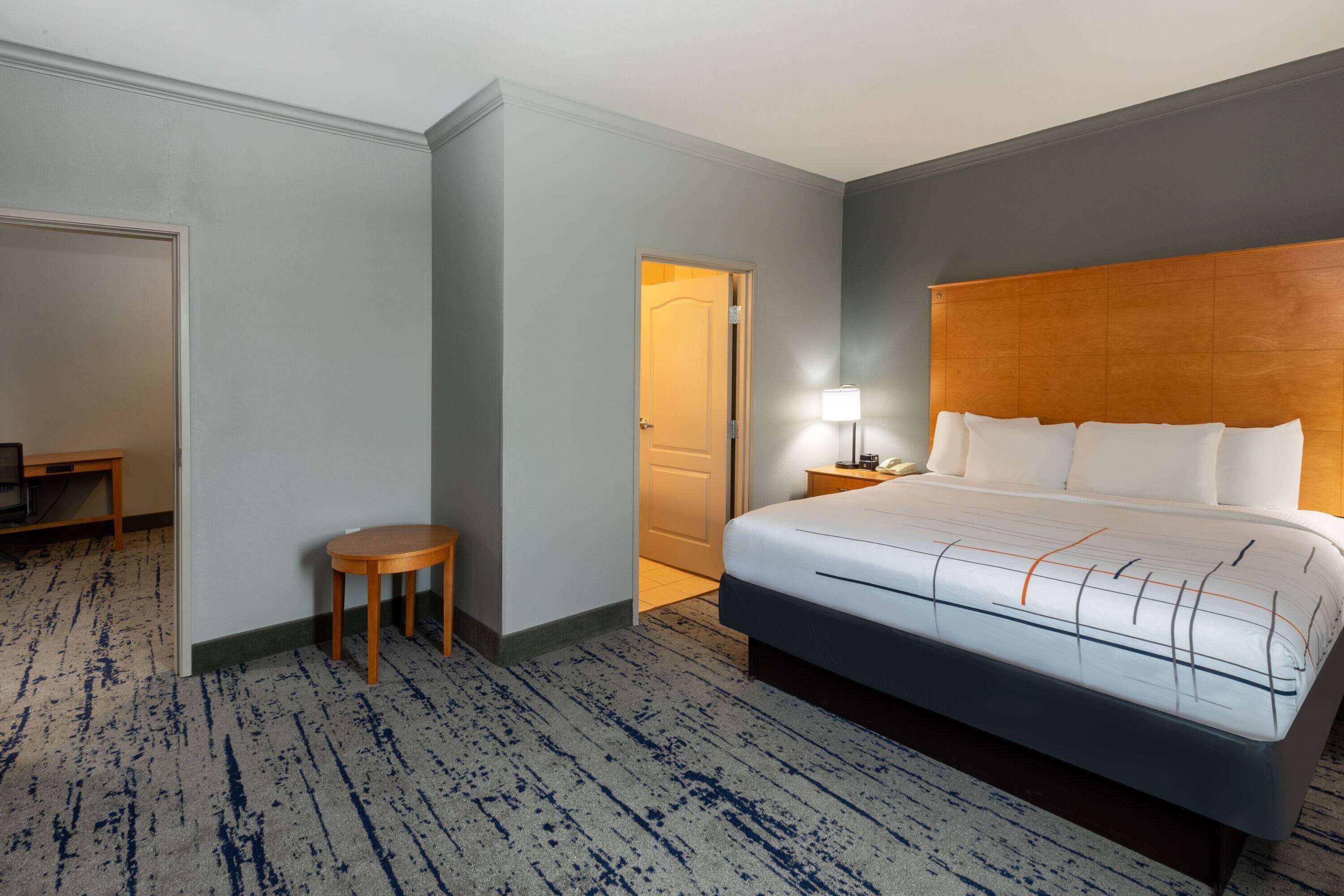 Ein Hotelzimmer mit einem großen Bett, einem kleinen Tisch, einem Nachttisch mit Lampe und einer Tür zu einem weiteren Raum.
