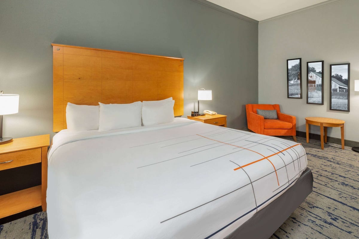 A hotel room with a large bed, a wooden headboard, a nightstand with a lamp, an orange chair, a small table, and three framed pictures on the wall.
