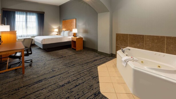 A hotel room with a large bed, a desk with a chair, a desk lamp, a nightstand with a lamp, and a bathtub. - La Quinta Inn & Suites by Wyndham Bridgeport (Bridgeport)