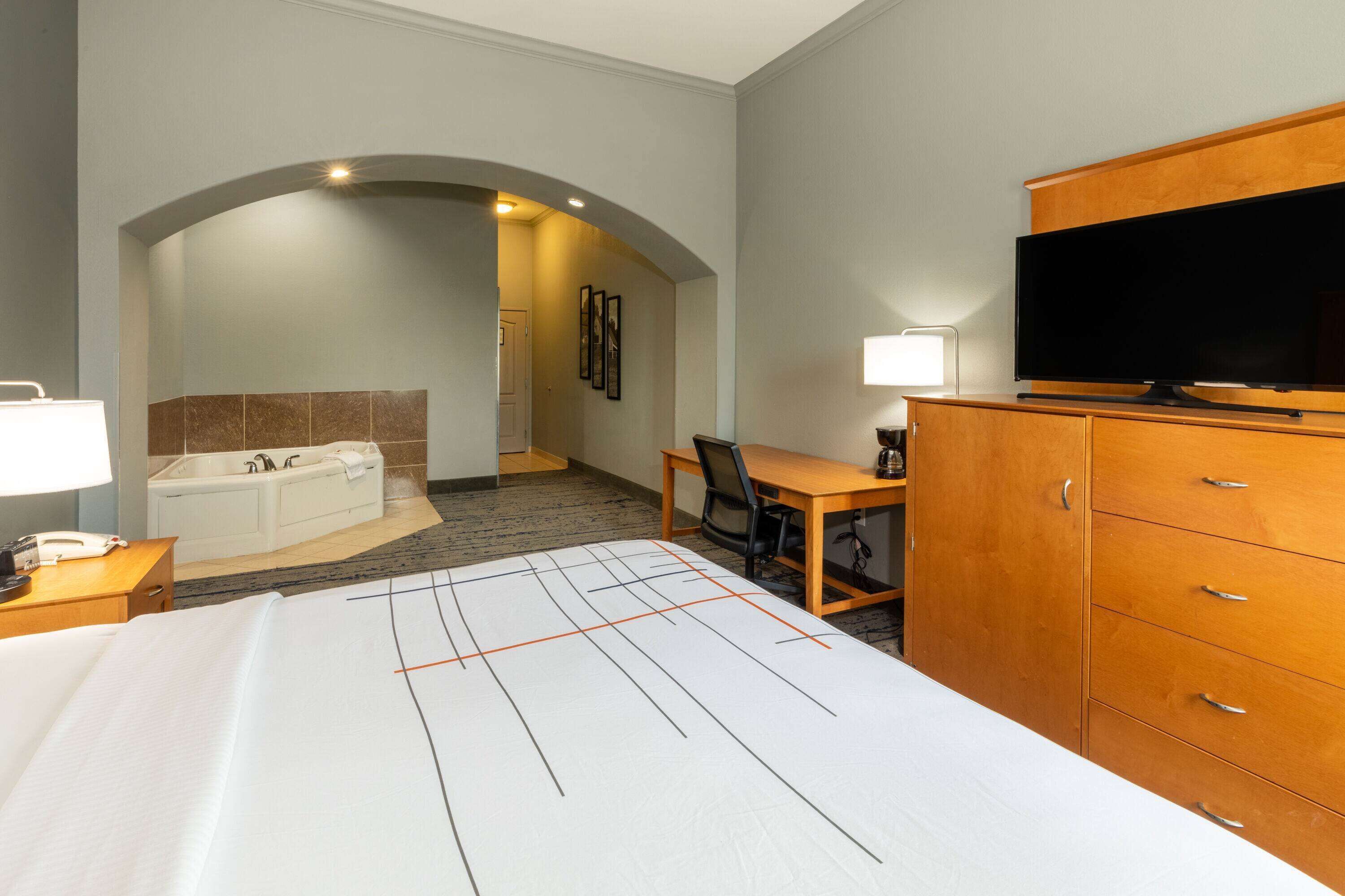 A hotel room with a bed, a desk, a television, a cabinet, a bathroom with a bathtub, and an arched doorway.
