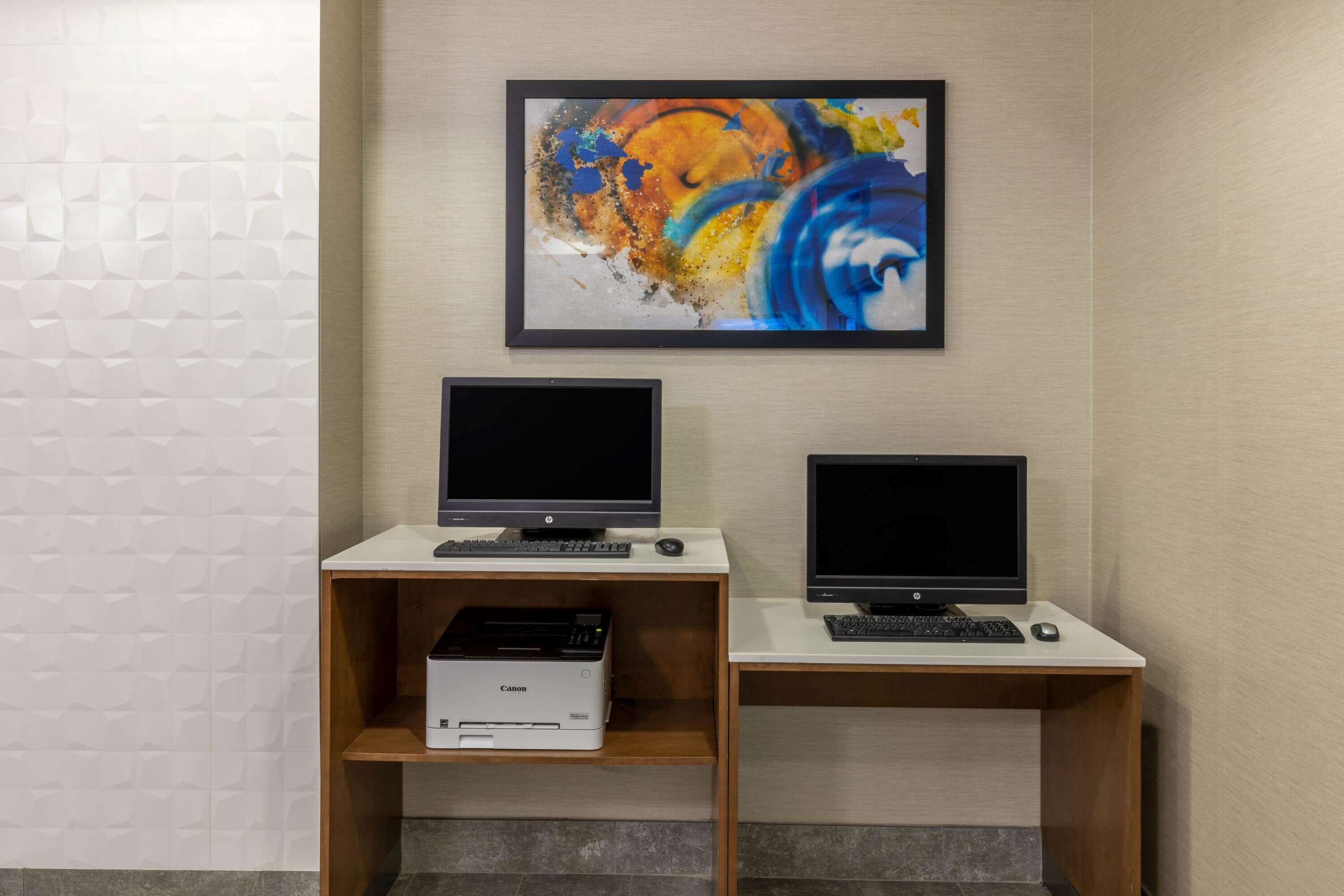Zwei Desktop-Computer an separaten Schreibtischen, ein Drucker links, ein gerahmtes abstraktes Gemälde über dem linken Schreibtisch und eine strukturierte Wand links.