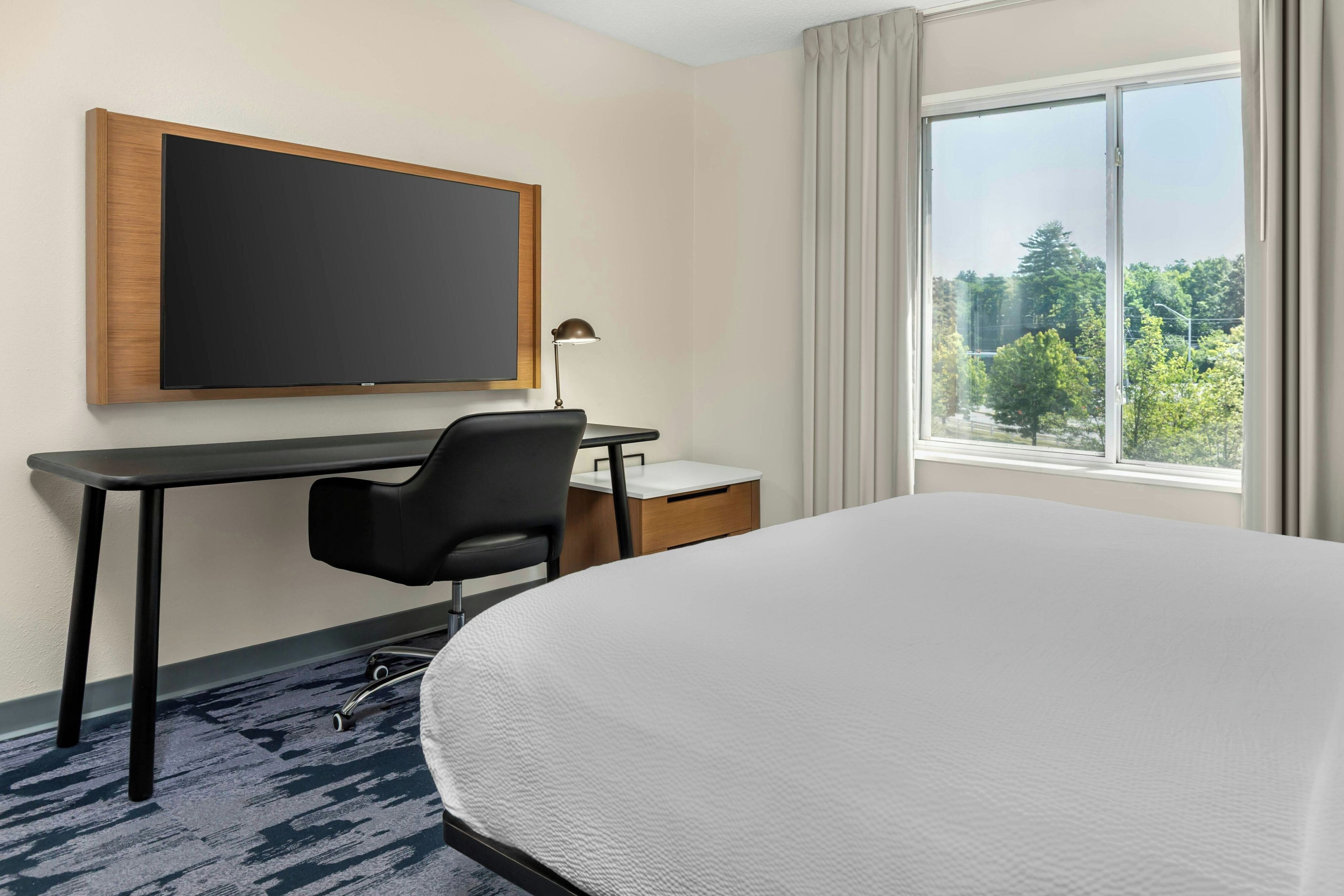 Habitación de hotel con cama, escritorio, silla, una pizarra y ventana con vista a árboles.