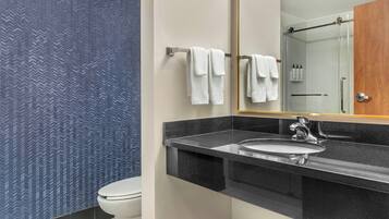 A modern bathroom with a black countertop, a white sink, and a mirror above it. There is a toilet to the left and a shower area to the right.