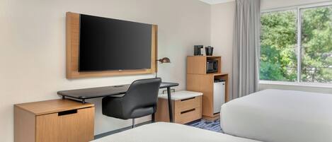 A hotel room with a bed, a desk with a chair, a television, a coffee maker, and a window with a view of trees.