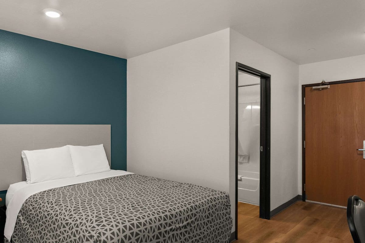 Un dormitorio con cama, mesita de noche, puerta de madera y baño visible a través de un acceso abierto.