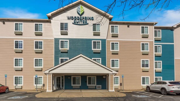 A multi-story hotel with a central entrance, multiple windows, and a parking lot with cars. - WoodSpring Suites Evansville East (Evansville)