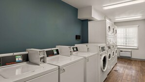 A laundry facility with multiple washing machines and dryers, a blue wall, and a window with blinds. - WoodSpring Suites Evansville East (Evansville)