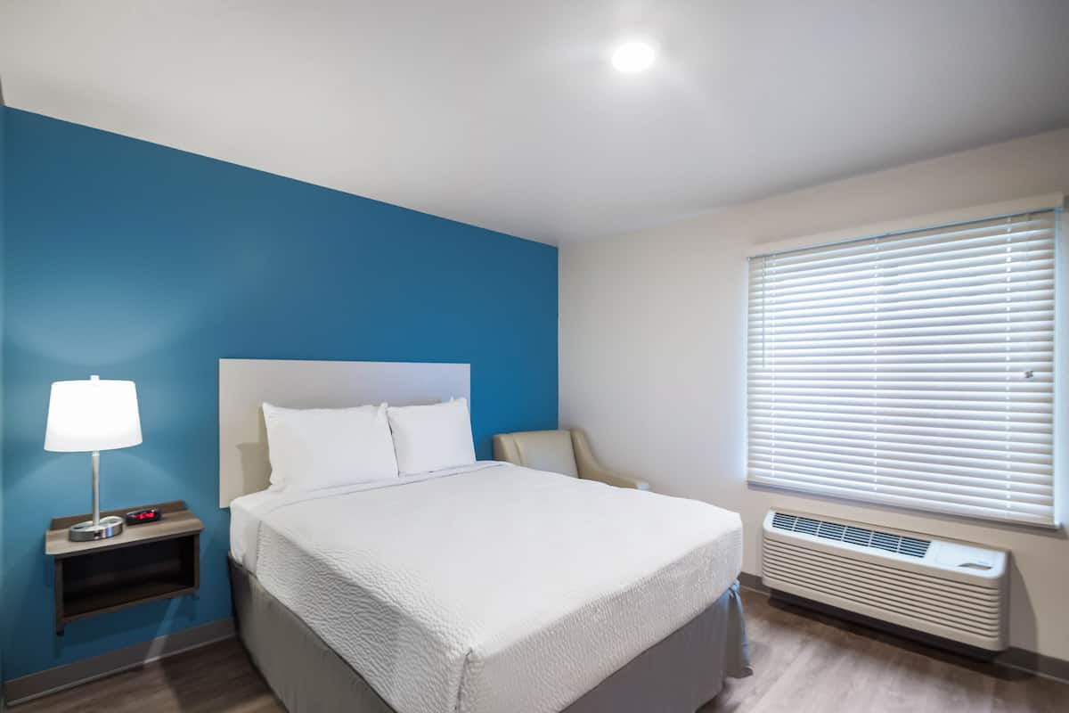 Habitación de hotel con cama, mesita de noche con lámpara, silla, ventana con persianas y calentador en la pared.