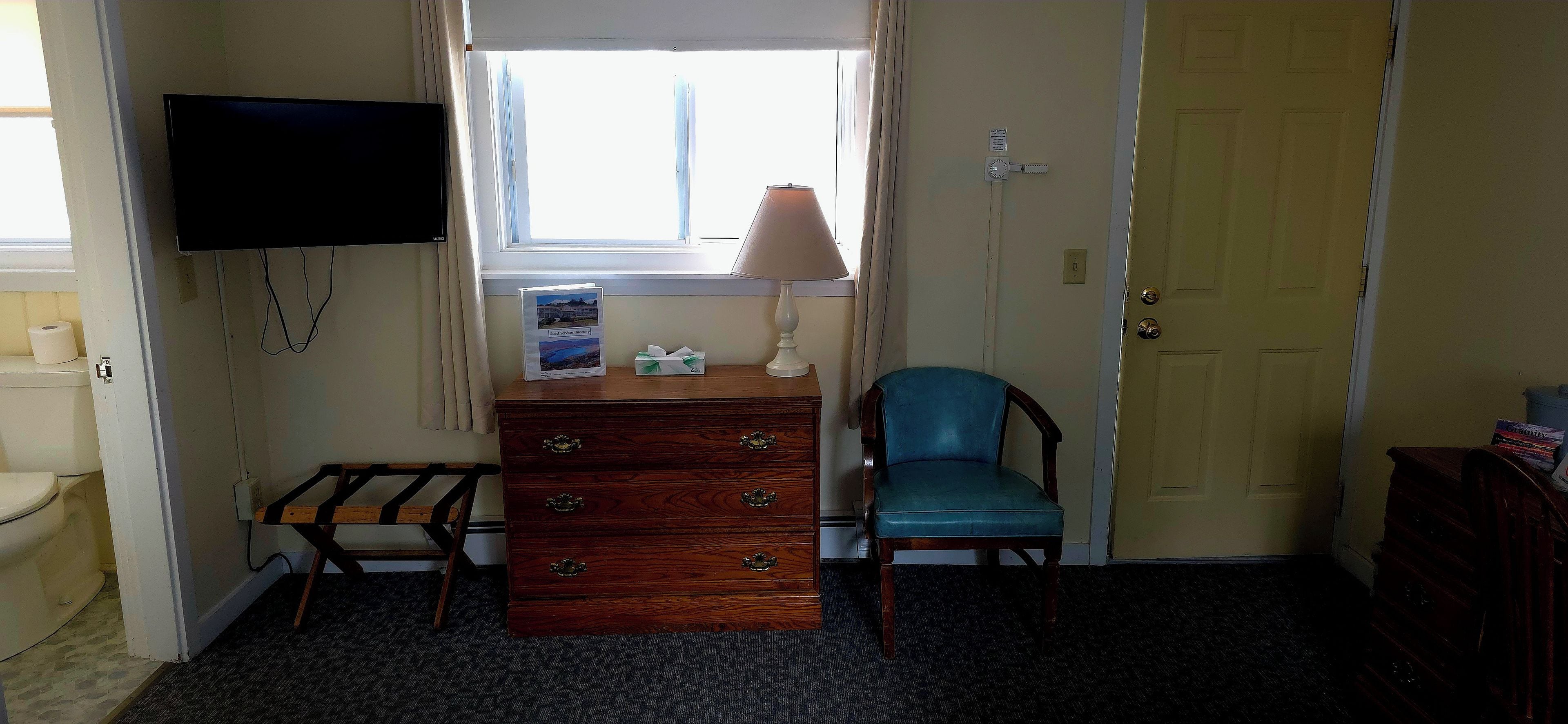 A room with a wooden dresser, a blue chair, a television mounted on the wall, and a window with curtains.