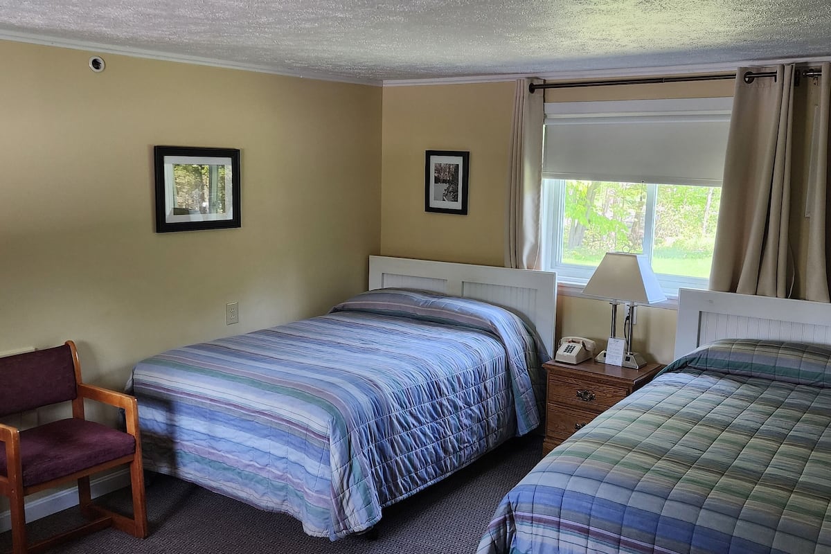 A hotel room with two beds, a chair, a nightstand with a lamp, and a window with curtains.