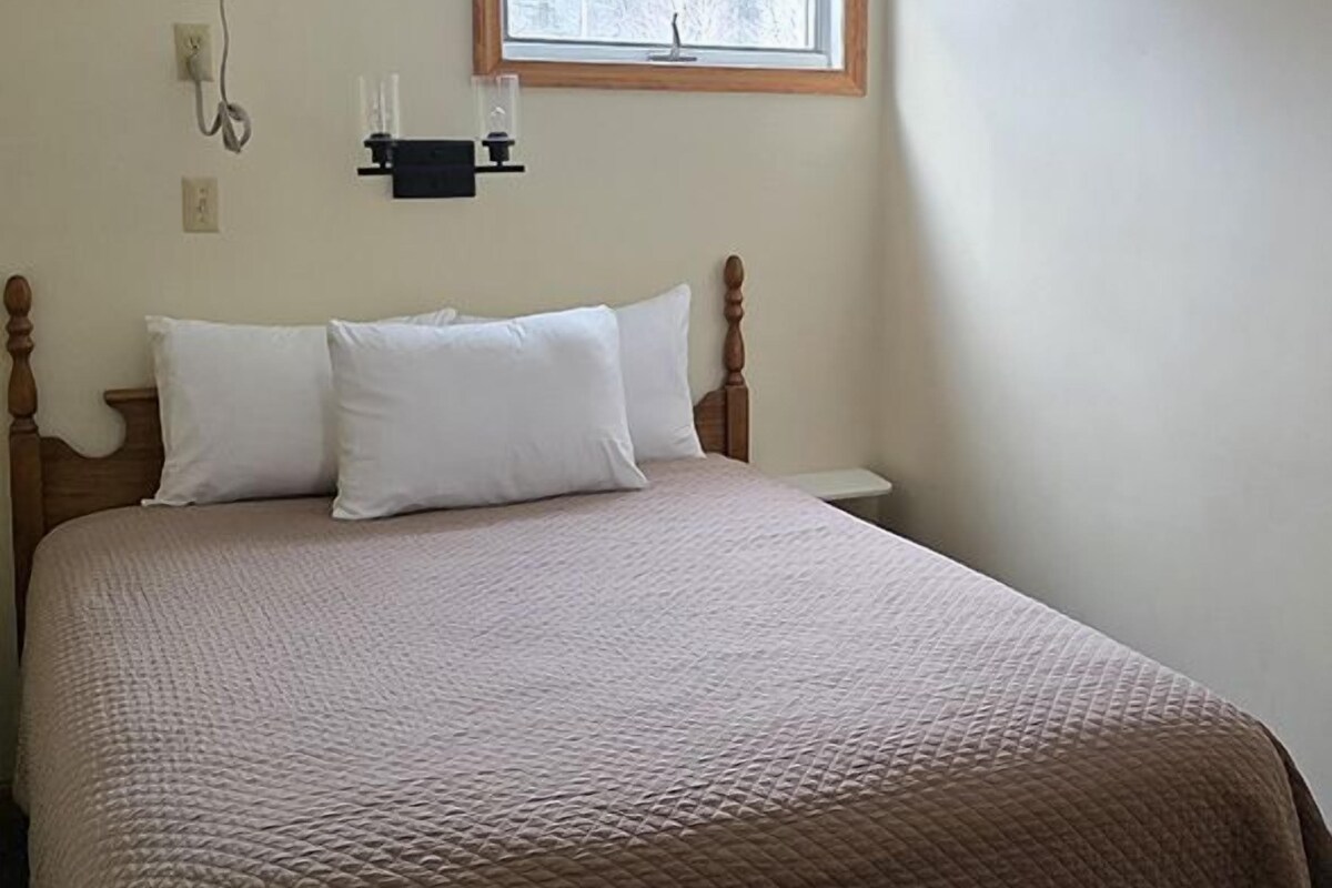 A bedroom with a bed, a window, a wall-mounted phone, and a light switch.