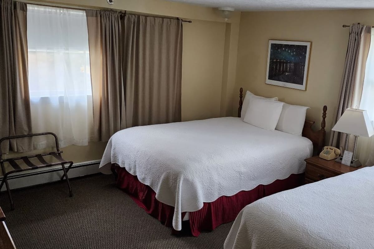 A hotel room with two beds, a bedside table with a lamp, a chair, and a painting on the wall.