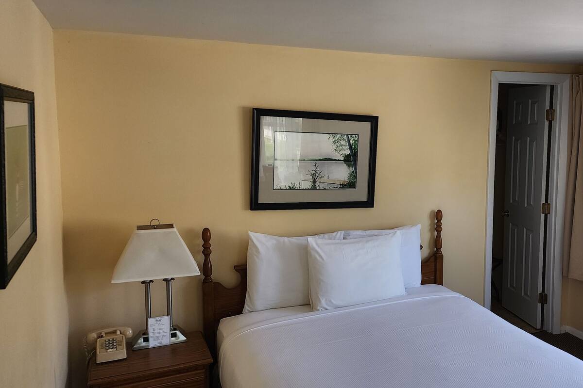 A hotel room with a bed, a bedside lamp, a telephone, a framed picture on the wall, and a window with curtains.