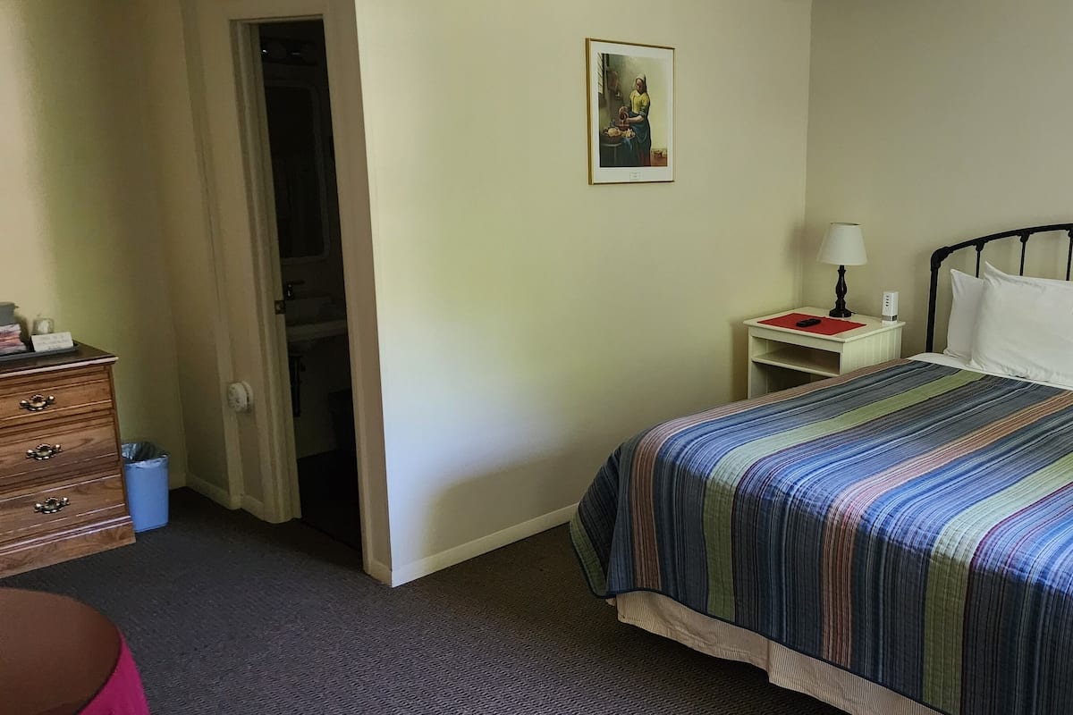 A hotel room with a bed, a bedside table with a lamp, a dresser, and a door leading to another room.