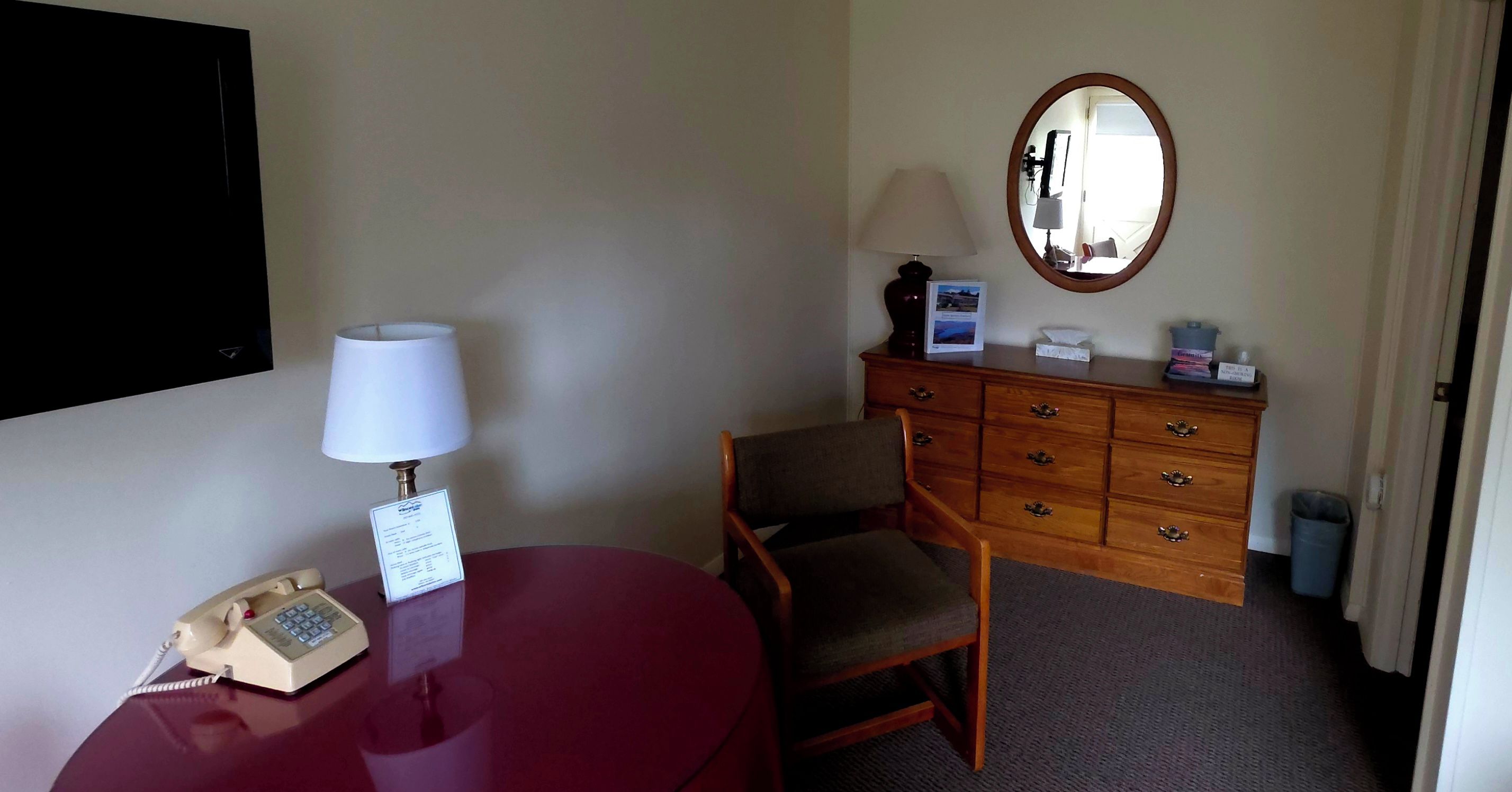 A hotel room with a desk, a telephone, a chair, a dresser, a mirror, and a carpeted floor.