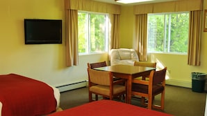 Quarto de hotel com duas camas, uma mesa de jantar e uma cadeira. Há janelas com cortinas, uma televisão na parede e uma lixeira.
