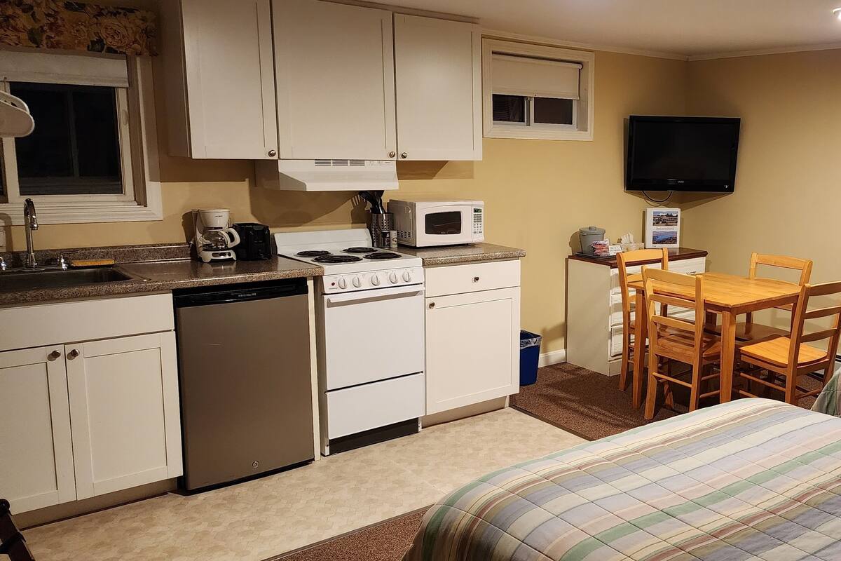 A compact kitchen with a stove, microwave, and sink. There is a dining table with chairs and two beds with striped bedding.