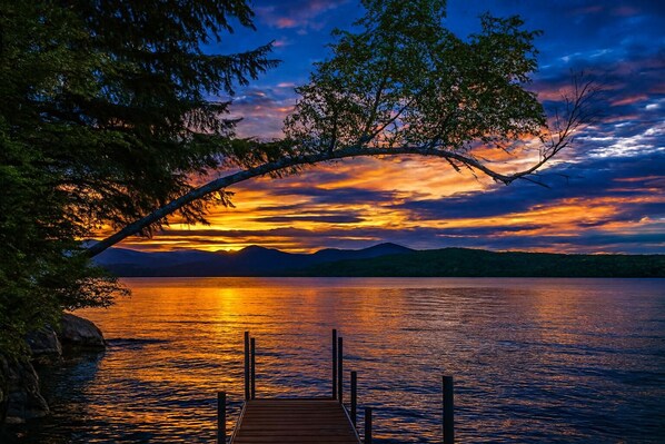 A wooden dock extending into calm water, surrounded by trees and mountains in the background.