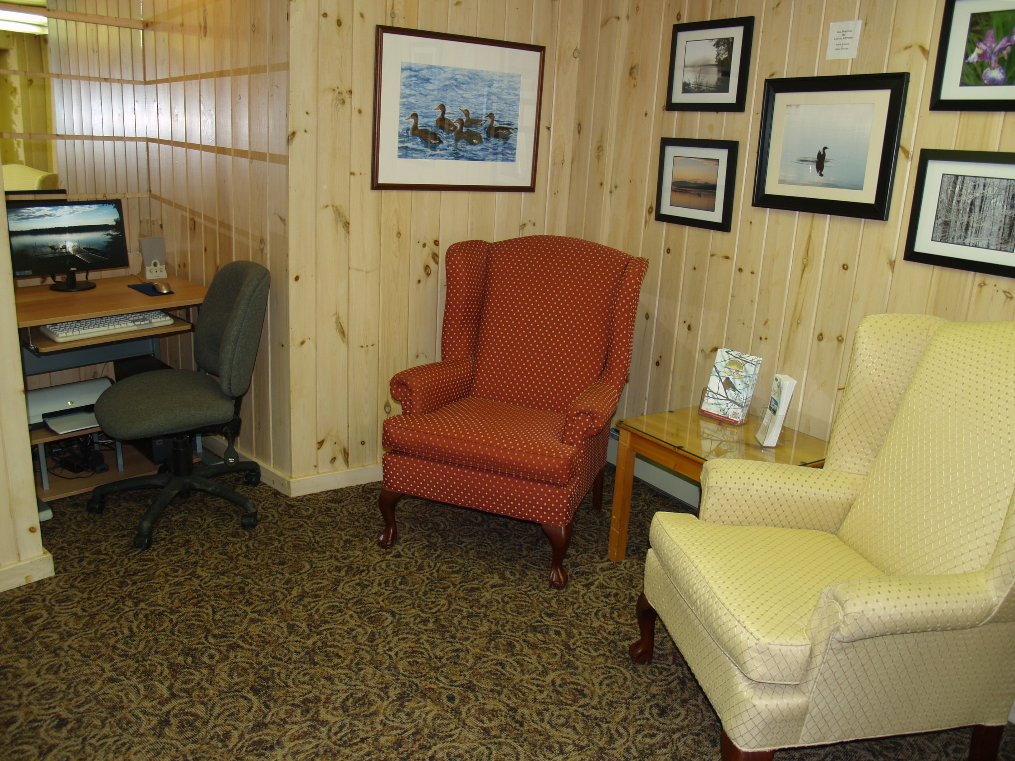 A room with a wooden paneled wall, a desk with a computer, two chairs, and framed pictures on the wall.