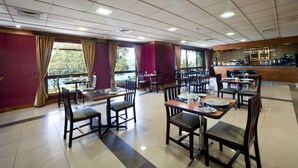 Un restaurante con varias mesas y sillas, una zona de bar y amplios ventanales con cortinas.