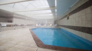 Una piscina interior con techo de vidrio transparente, paredes de azulejos y sillones de descanso alrededor.