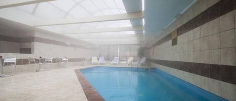 An indoor swimming pool with clear glass ceiling, tiled walls, and lounge chairs around the pool.