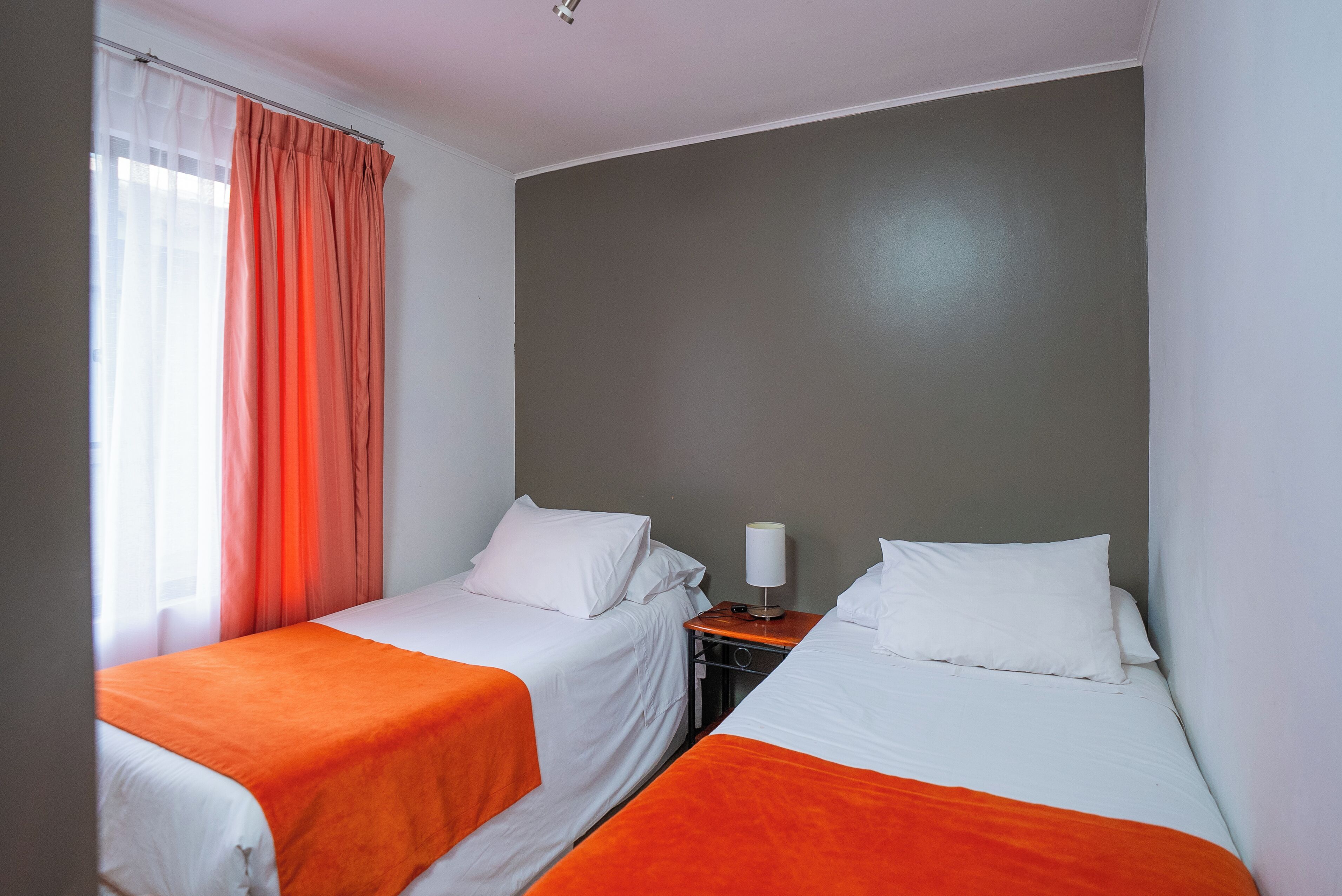 Quarto de hotel com duas camas individuais, cada uma com roupa de cama branca e mantas laranja. Há uma mesa de cabeceira com uma luminária entre as camas.