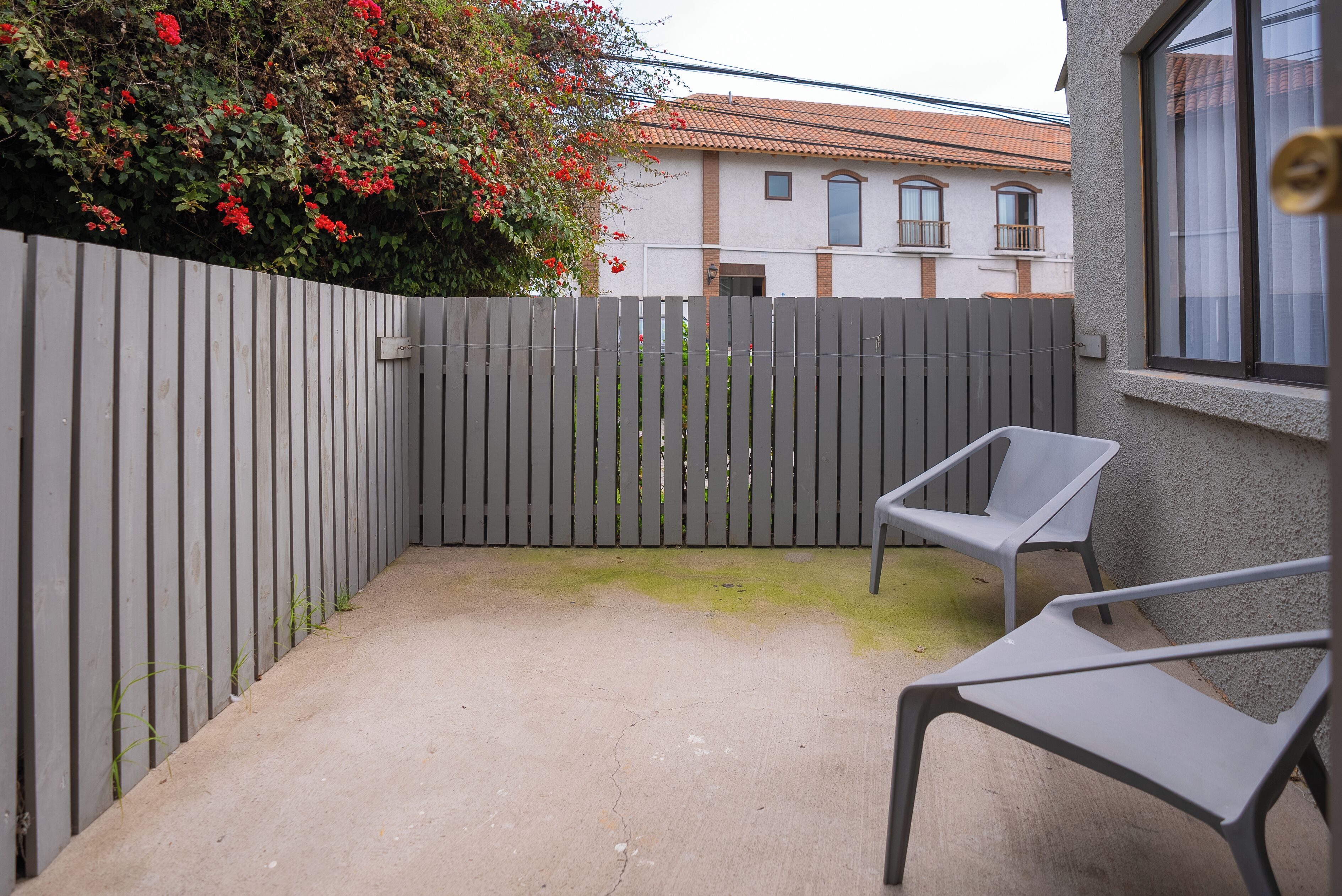 A patio with a grey fence, two grey chairs, and a building with a tiled roof in the background.