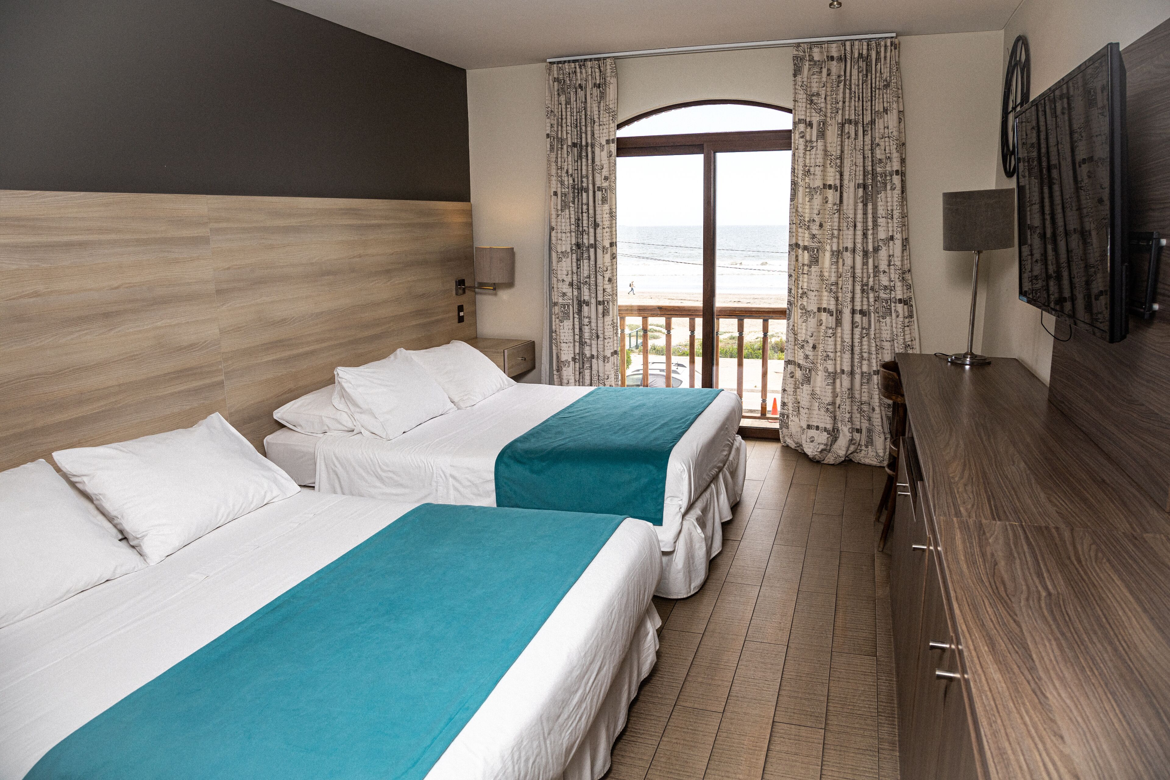 Une chambre d’hôtel avec deux lits, une tête de lit en bois, un balcon avec des rideaux, une télévision et un bureau.