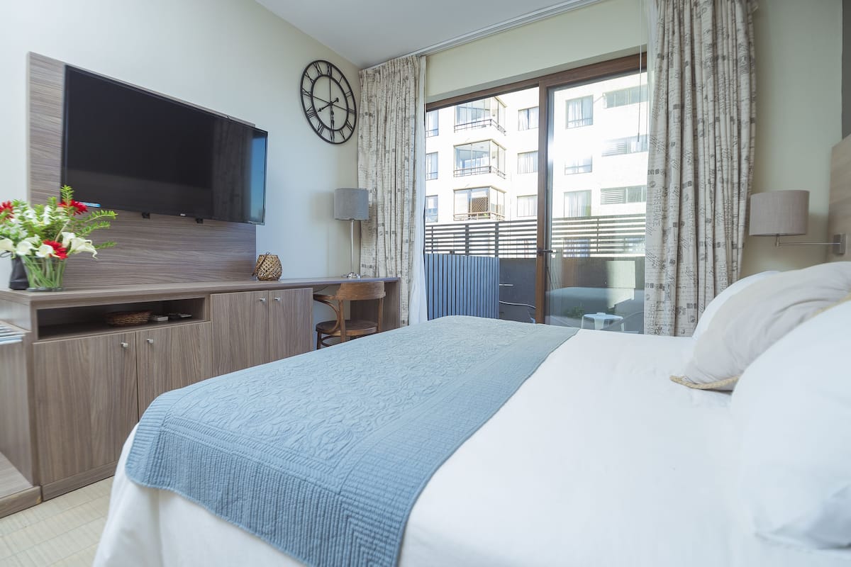Habitación de hotel con una cama grande, un televisor de pantalla plana en la pared, un escritorio con lámpara y vistas al balcón exterior.