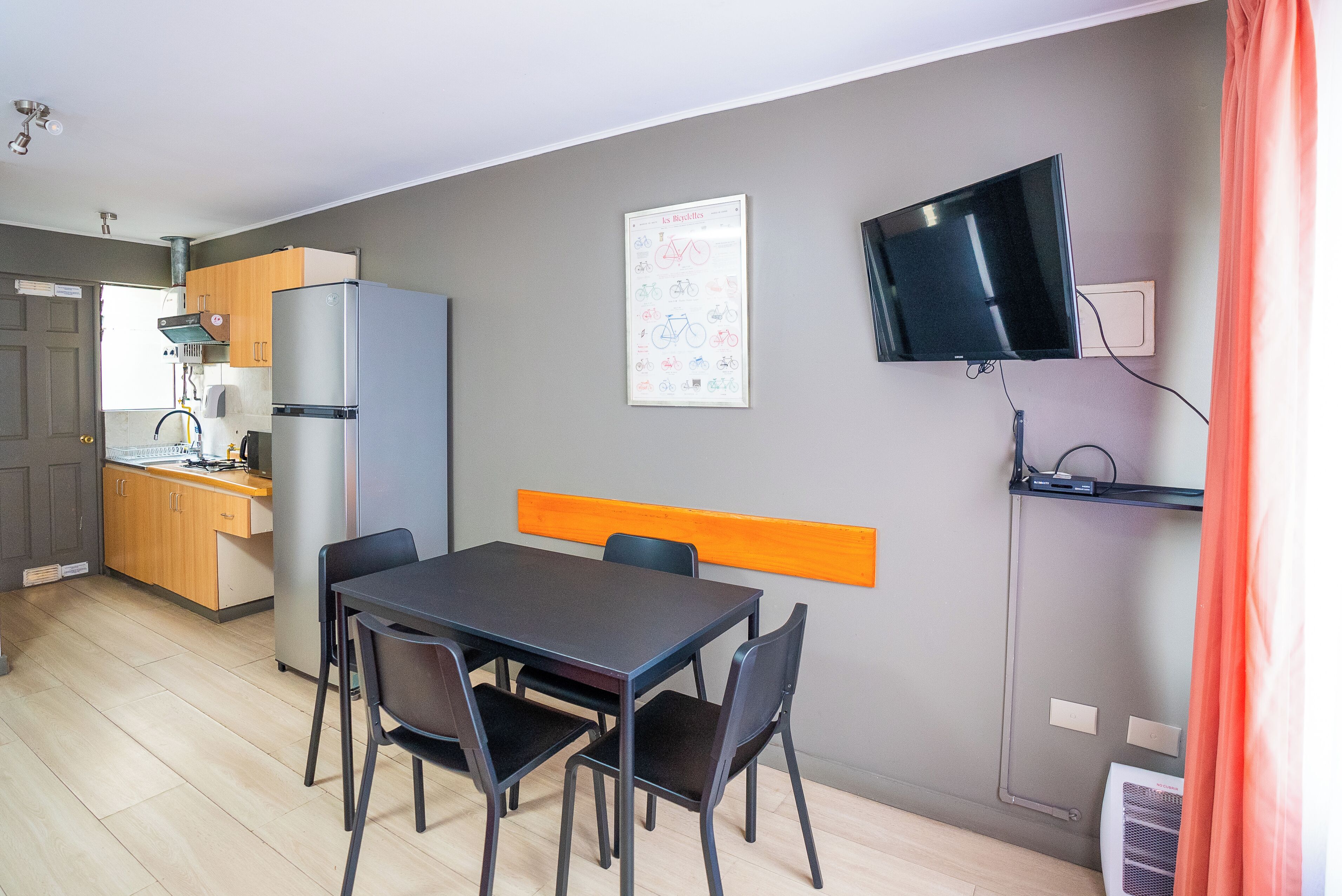 Una cocina compacta equipada con refrigerador, estufa y lavaplatos. Cuenta con un comedor con mesa y sillas, y un televisor montado en la pared.