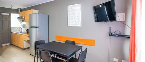 Una cocina compacta equipada con refrigerador, estufa y lavaplatos. Cuenta con un comedor con mesa y sillas, y un televisor montado en la pared.