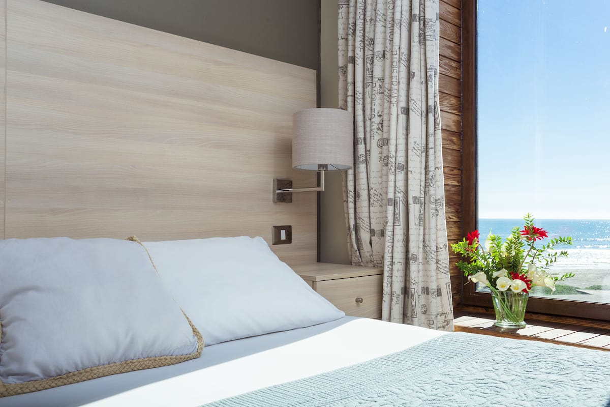 Un cuarto de hotel con una cama, una mesita de noche, una lámpara, un florero con flores y vista al océano.