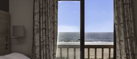 A room with a bed, a nightstand, a lamp, and a view of the ocean through a large window.