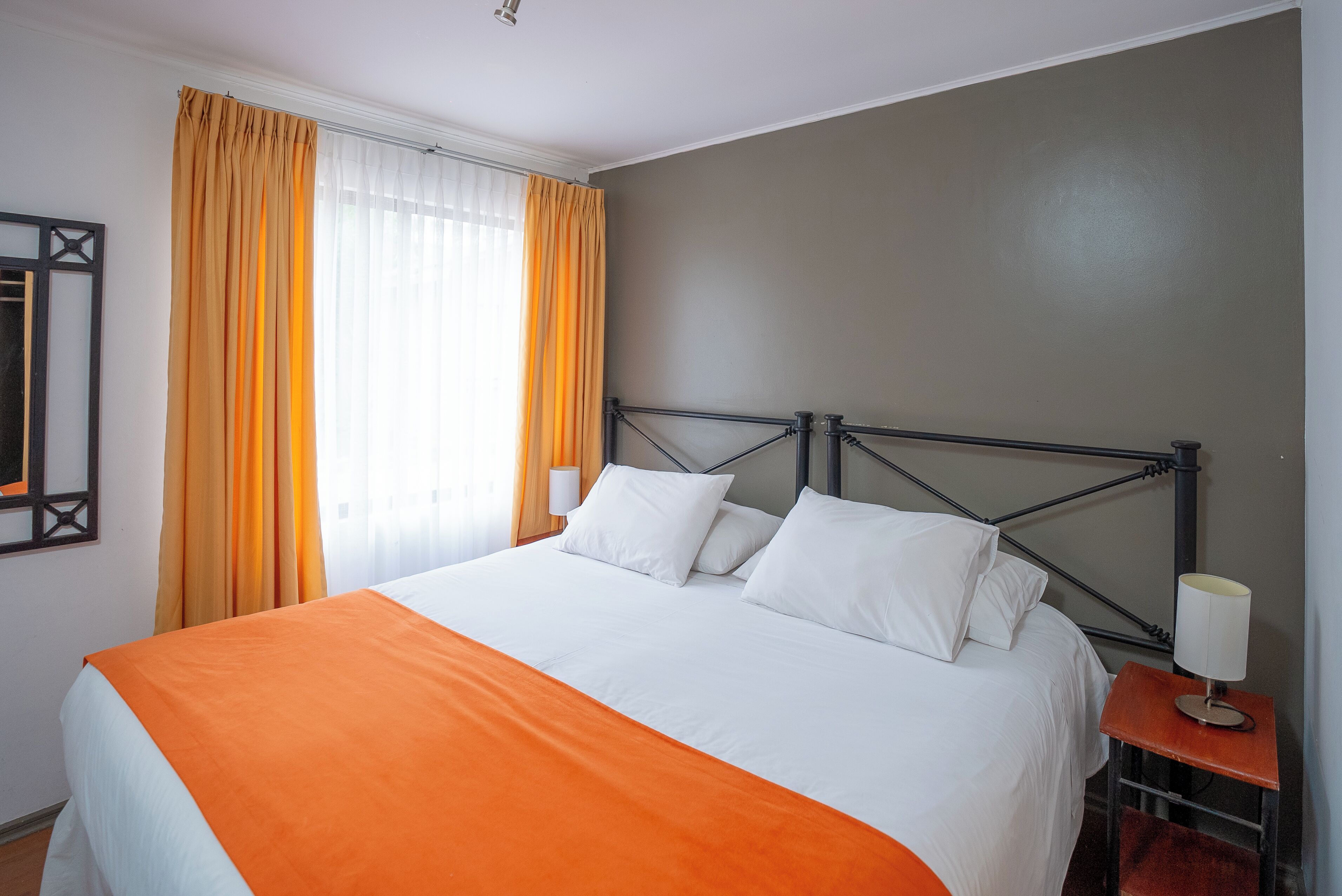 Una cama bien hecha con ropa de cama blanca y naranja, un cabecero de metal y una mesita de noche con una lámpara.